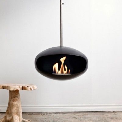 A sleek, modern, hanging fireplace with a rounded black exterior and a visible flame burning inside. The minimalist design is complemented by a natural wood table beside it, and the clean, white walls and floor provide a simple, neutral backdrop.